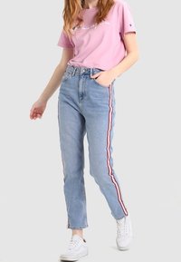 Jeans de color azul claro con rayas laterales rojas y blancas, de cintura alta y con un corte recto, combinados con una camiseta rosa de manga corta y zapatillas blancas.