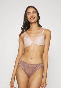 Smaidīga sieviete ar gariem tumšiem matiem, valkājot gaiši rozā mežģīņu krūšturi un violetas mežģīņu biksītes uz vienkārša gaiša fona.