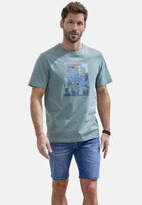 Helles grünes Baumwoll-T-Shirt mit einem bunten Grafikdruck auf der Vorderseite, kombiniert mit Jeansshorts. Lässiges Design mit kurzen Ärmeln.