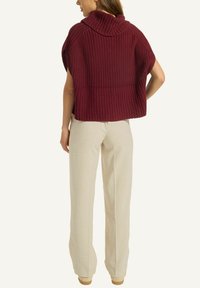 Eine Frau trägt einen ärmellosen, grob gestrickten kastanienbraunen Pullover mit einem Schalkragen und lockeren, beigefarbenen Hosen und steht mit den Händen hinter dem Rücken.