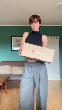 Kartonasti zaboj s logotipom Zalando, ki ga drži oseba v črnem brezrokavniku in s sivo širokimi hlačami v notranjem prostoru.