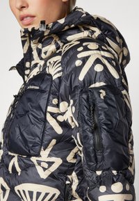 Casaco acolchoado com fundo azul-marinho e padrões abstratos bege. Possui capuz, bolsos com fecho éclair e textura acolchoada. Material leve.