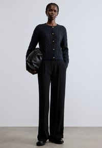 Καφέ πλεκτή ζακέτα με χρυσά κουμπιά, συνδυασμένη με μαύρο παντελόνι με φαρδιά πόδια και μαλακή μαύρη τσάντα, σε ουδέτερο φόντο.