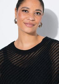 Svart stickad tröja med en lös, öppen väv och vid halsringning. Modellen visas med silverfärgade ringörhängen som framhäver en jämn hudtextur.