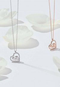 To hjerteformede halskæder: en sølvfarvet med en tekstureret kant og en rosaguld, begge med dekorative detaljer og hængende kæder.