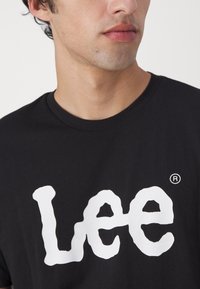 Melna kokvilnas T-krekls ar lielu, baltu, stilizētu "Lee" logo, apaļu kakla izgriezumu, īsām piedurknēm un reģistrētu preču zīmi.