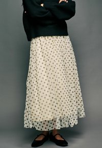 Pull en tricot noir, jupe maxi en tulle crème à pois, longueur cheville, texture fluide. Ballerines noires avec détail de sangle. Fond gris.