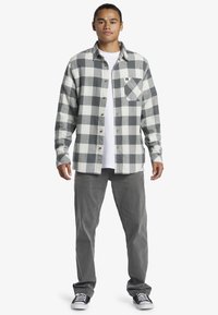 Camicia di flanella con motivo a quadri grigi e bianchi, design con bottoni e taschino sul petto, indossata sopra una t-shirt bianca e abbinata a pantaloni grigi.