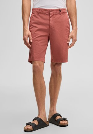 Mann trägt rostfarbene knielange Shorts, weißes T-Shirt und schwarze Sandalen mit Doppelriemen, steht vor einem einfarbigen Hintergrund.