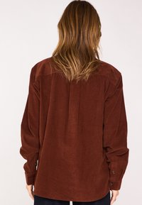 Camicia di velluto a costine marrone con una vestibilità comoda, polsini con bottoni e una pince centrale sul retro, caratterizzata da un tessuto morbido e testurizzato.