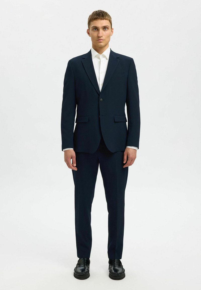 Marinblå skräddarsydd kostym med enbladsblazer, vit skjorta och svarta skor. Slät textur med slim-fit design och rena linjer.