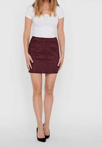 Frau in einem weißen, kurzärmligen, figurbetonten Oberteil, bordeauxrotem Wildleder-Minirock und schwarzen High Heels, die vor einem schlichten Hintergrund steht.