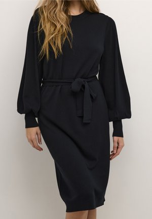 Femme portant une robe midi noire avec des manches bouffantes et une ceinture nouée à la taille, debout devant un fond clair uni.