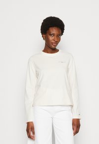 Top a maniche lunghe color crema con scollatura rotonda, caratterizzato da una piccola stampa del logo sul petto e una vestibilità comoda. Indossato con pantaloni bianchi.