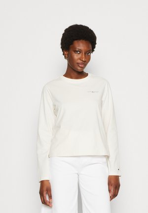 Top a maniche lunghe color crema con scollatura rotonda, caratterizzato da una piccola stampa del logo sul petto e una vestibilità comoda. Indossato con pantaloni bianchi.