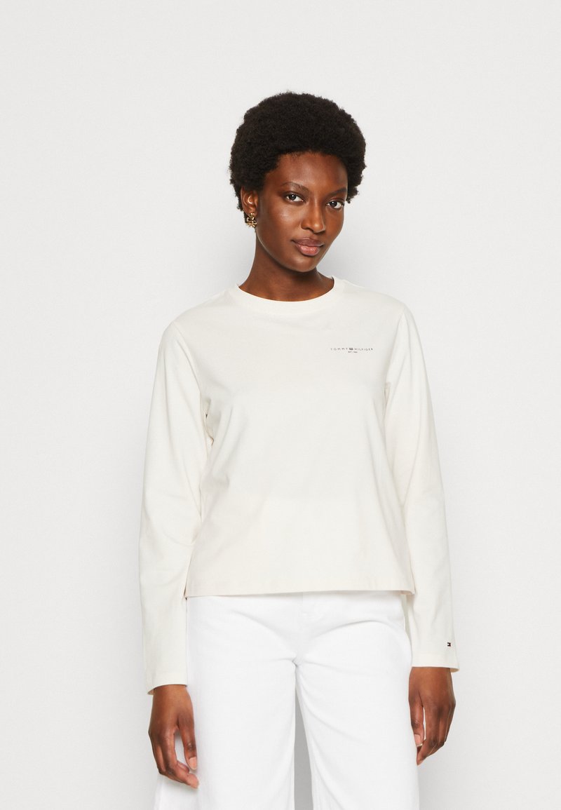 Top a maniche lunghe color crema con scollatura rotonda, caratterizzato da una piccola stampa del logo sul petto e una vestibilità comoda. Indossato con pantaloni bianchi.