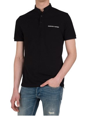 Mężczyzna ubrany w czarną koszulkę typu polo z krótkim rękawem z logo "KOOPLES X SPORT", w połączeniu z niebieskimi, przetartymi dżinsami, z ręką w kieszeni.