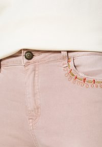 Jeans de un suave color rosa claro con una textura lisa, presentando un botón en tono dorado con diseño de rayos de sol y acentos bordados en rojo y amarillo a lo largo del bolsillo.