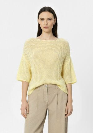 Junge Frau mit glattem dunklem Haar, die einen locker sitzenden, hellgelben Strickpullover und beige hoch taillierte Hosen trägt, stehend vor einfarbigem Hintergrund.