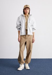 Grå zip-up hettegenser, beige skjorte med knapper, brune cargo-bukser og hvite høye sneakers. Enkel caps med "LA"-logo.