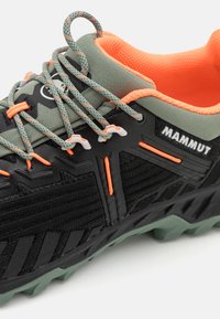 Scarpa da trail nera e robusta con suola verde, dettagli grigi e arancioni, tomaia sintetica testurizzata e punta rinforzata. Logo del brand visibile.