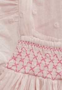 Helles rosa Gewebe mit strukturierten Punkten und pinken Smock-Details. Verfügt über gerüschte Kanten und ein gerafftes Design für zusätzliches Volumen.