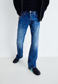 Jeans de denim azul con corte recto, que presentan un acabado desgastado y un diseño clásico de cinco bolsillos. Combinados con zapatillas negras y una chaqueta negra.