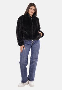 Giacca in pelliccia sintetica nera con colletto alto e zip frontale, abbinata a jeans di denim blu e sneakers bianche. Texture liscia su tutta la superficie.
