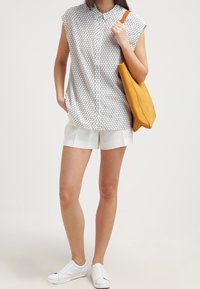 Kvinna klädd i vita shorts, ärmlös blus med vita prickar, vita slip-on skor, som håller en senapsgul axelväska.