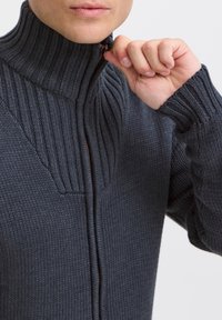 Kolgrå stickad zip-up-tröja med hög krage, ribbad textur och en partiell dragkedja. Noterbara detaljer inkluderar kontrasterande stickmönster.