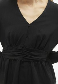Blusa nera con scollatura a V, con bottoni frontali, vita arricciata e una texture morbida e fluida. Il tessuto sembra leggero.