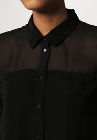 Black button-up shirt with a transparent upper panel, featuring a classic collar and two visible buttons on the front, smooth texture.
