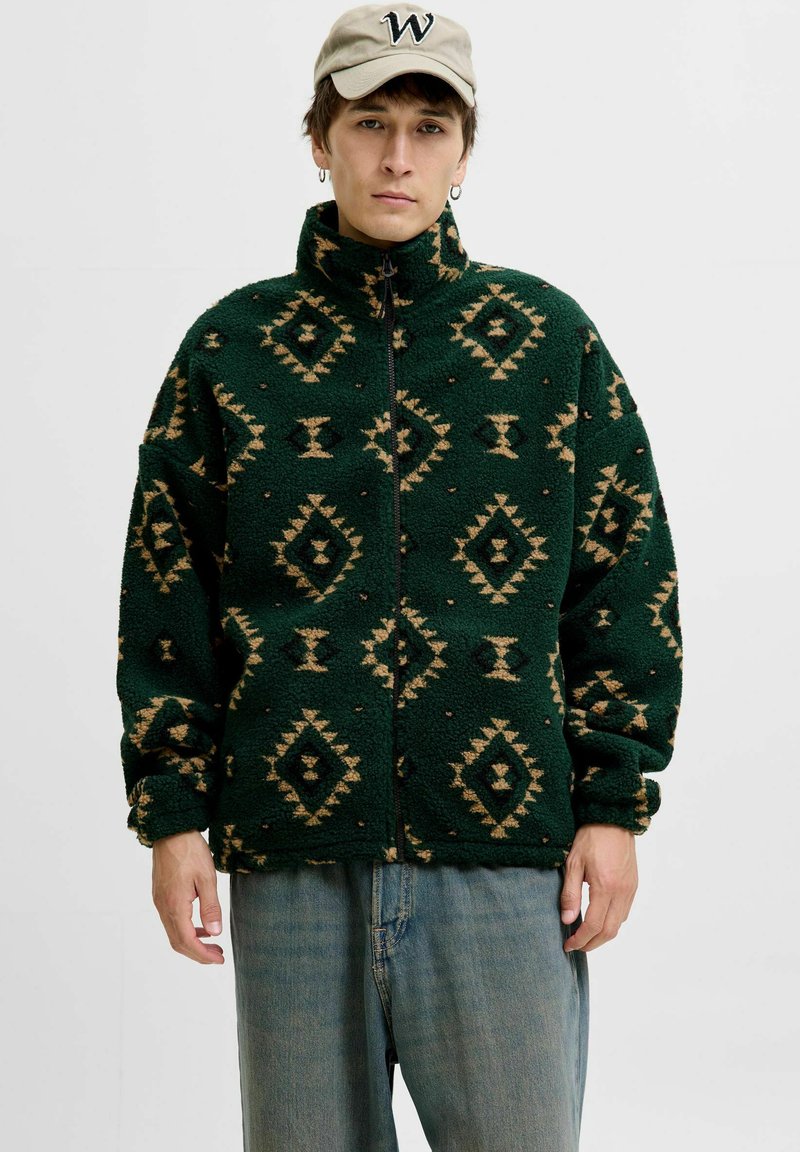 Grüne Fleecejacke mit geometrischen hellbraunen Mustern, hohem Kragen, durchgehendem Reißverschluss und übergroßen Ärmeln. Kombiniert mit hellblauen Jeans.