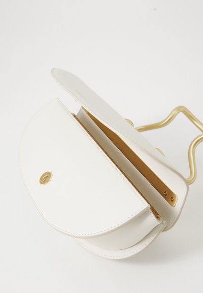 Valkoinen nahkainen puolikuun muotoinen crossbody-laukku, jossa on kultainen magneettisuljin ja muotoiltu kultainen kahva, avattuna valkoista taustaa vasten.
