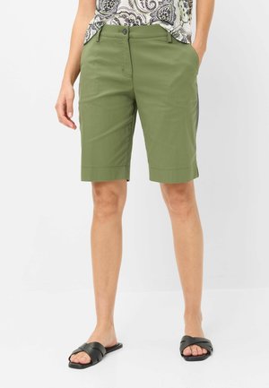 Frau trägt olivgrüne kniehohe Shorts, ein gemustertes Oberteil und schwarze flache Sandalen, steht mit Händen in den Taschen.