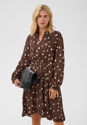 Femme aux cheveux blonds mi-longs porte une robe marron à pois à manches longues et tient une grande pochette noire devant un fond uni.