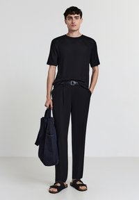 Uomo che indossa una camicia nera a maniche corte con texture, pantaloni neri, cintura nera, sandali neri, mentre tiene una grande borsa a tracolla nera, in piedi contro uno sfondo neutro.