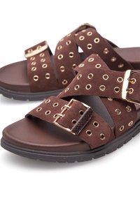 Sandali in suede marrone con fasce incrociate dotate di occhielli dorati e una fibbia metallica. Suola in gomma spessa con un pattern antiscivolo.