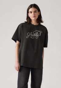 Camiseta negra tamaño grande hecha de algodón, con un gráfico en estilo script que dice "Howdy" en blanco y rojo en el frente. Textura suave, ajuste relajado.