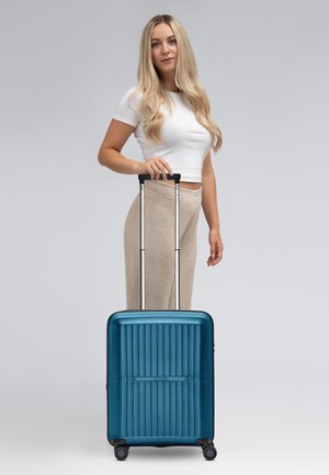 Valigia rigida di colore teal intenso, con design a righe verticali, maniglia per il trasporto e manico a trolley estensibile argento con 4 ruote nere.