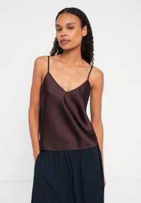 Kobieta w jedwabnym, ciemnobrązowym topie na cienkich ramiączkach i granatowej, plisowanej spódnicy, stojąca z jedną ręką w kieszeni na białym tle.