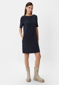Dunkelblaues Kurzarmkleid mit seitlichen Taschen, gerader Silhouette, Rundhalsausschnitt aus glattem Material. Ergänzt mit beigen kniehohen Stiefeln.