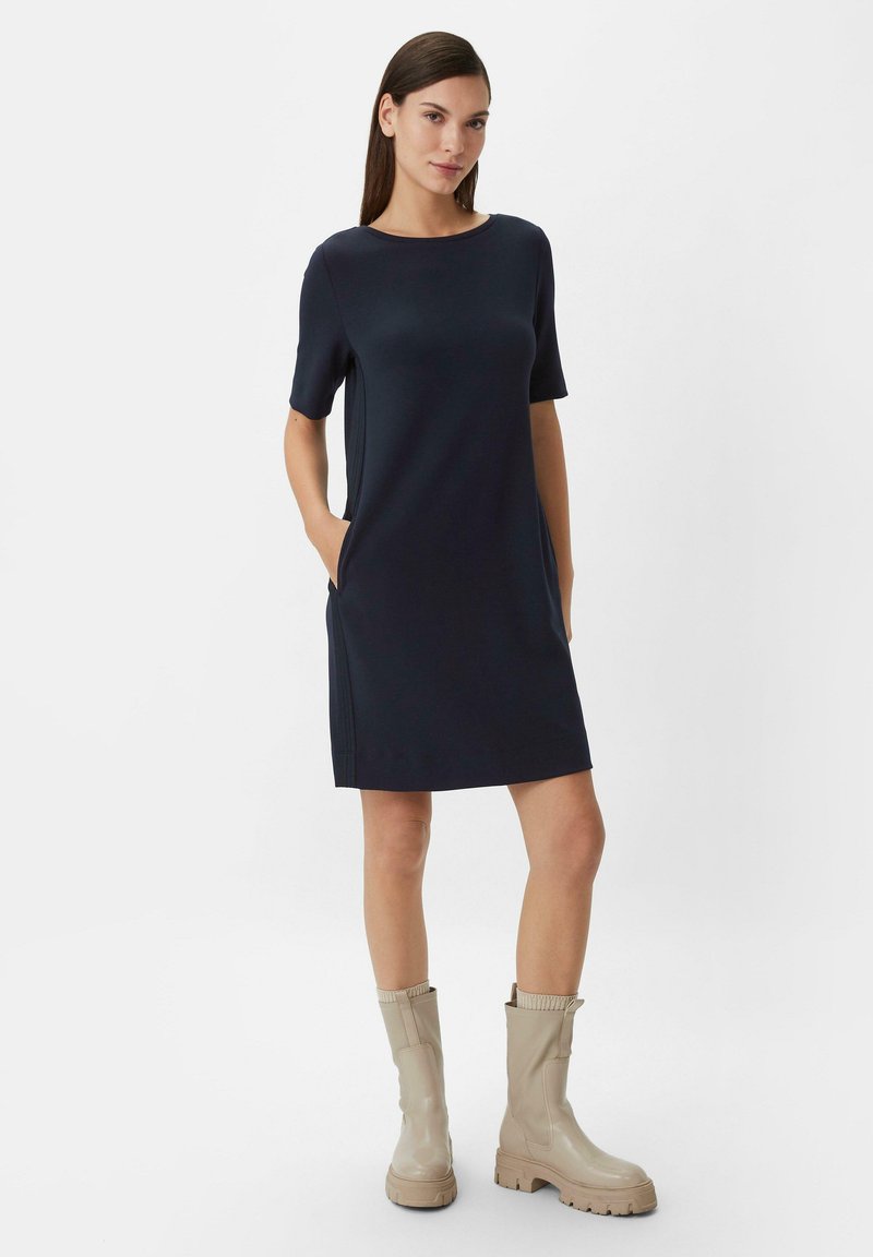Dunkelblaues Kurzarmkleid mit seitlichen Taschen, gerader Silhouette, Rundhalsausschnitt aus glattem Material. Ergänzt mit beigen kniehohen Stiefeln.