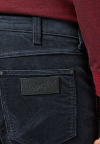 Donkerblauwe corduroy broek heeft een zwart leren Wrangler-logo patch op de achterzak, met subtiele stikdetails en een gestructureerd oppervlak.