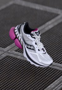 White athletic shoe with mesh upper, black accents, pink highlights, and a prominent cushioned sole design. Features lacing system.
