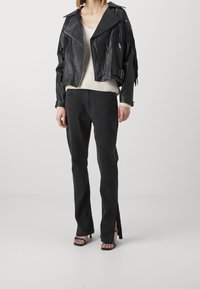 Zwarte leren cropped jas met franjes, gecombineerd met zwarte jeans met hoge taille en zijsplitsingen. Model draagt een crème kleurige gebreide top.