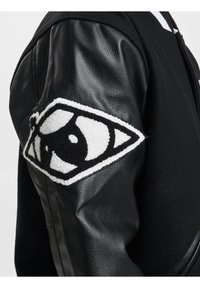 Veste noire avec un design en laine et cuir. Comprend un détail de patch brodé texturé en forme d'œil sur la manche gauche.