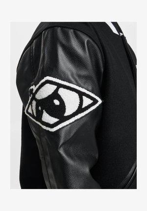 Musta takki, jossa on villa- ja nahkadesign. Sisältää teksturoidun, brodeeratun silmälaastarin vasemmassa hihassa.