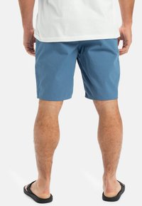 Blå bomullsshorts med en avslappnad design, med en lös passform och en fåll strax ovanför knäet. Bärs med svarta flip-flops.