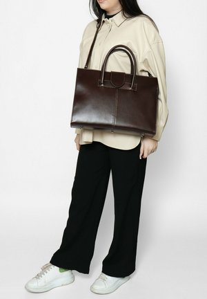 Borsa a tracolla in pelle marrone con due manici, design strutturato e cuciture visibili. Indossata da una persona con una maglietta neutra e pantaloni neri.